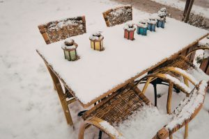 An outdoor cafe table and wicker chairs completely covered in a fresh layer of white snow. Five colorful vintage-style glass lanterns sit on the tabletop, partially buried in the snow, creating a quiet and cold winter scene.